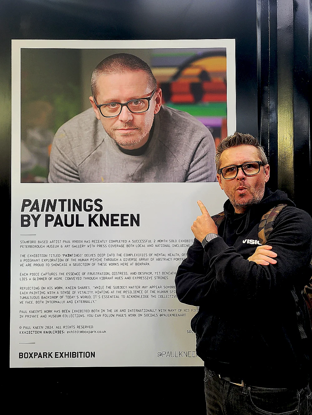 Artist Paul Kneen standing beside exhibition poster at BOXPARK Shoreditch